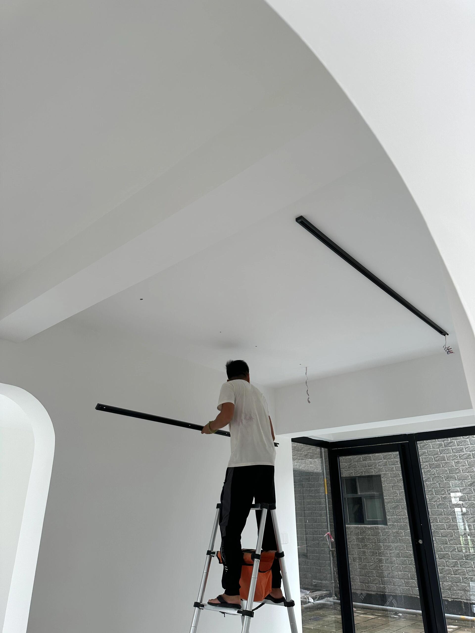 Professional electrician installing black surface-mounted track light on white ceiling - showing exposed track installation process