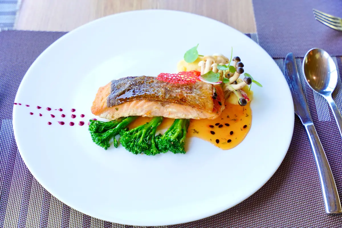 Gourmet salmon dish plated with greens, mushrooms and sauce under warm restaurant lighting, showing how cool ceramics and herbs read at 2700K
