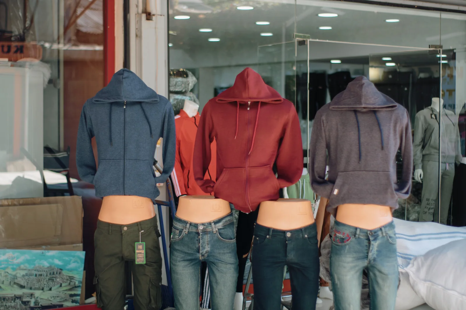 Retail storefront window display with hooded sweatshirts and jeans on mannequins lit by track heads against a dark interior