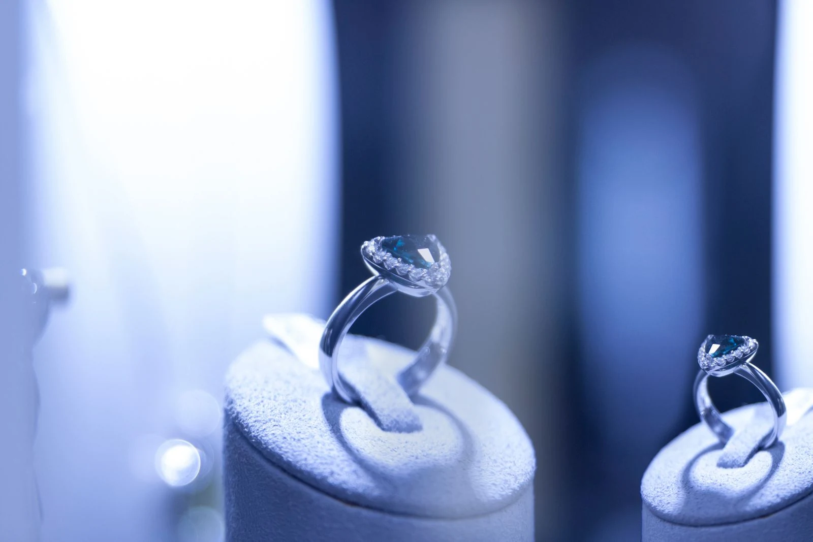 Two solitaire diamond rings on pale display plinths under a strong narrow spotlight, showing the tight pool a jewelry store hero piece needs