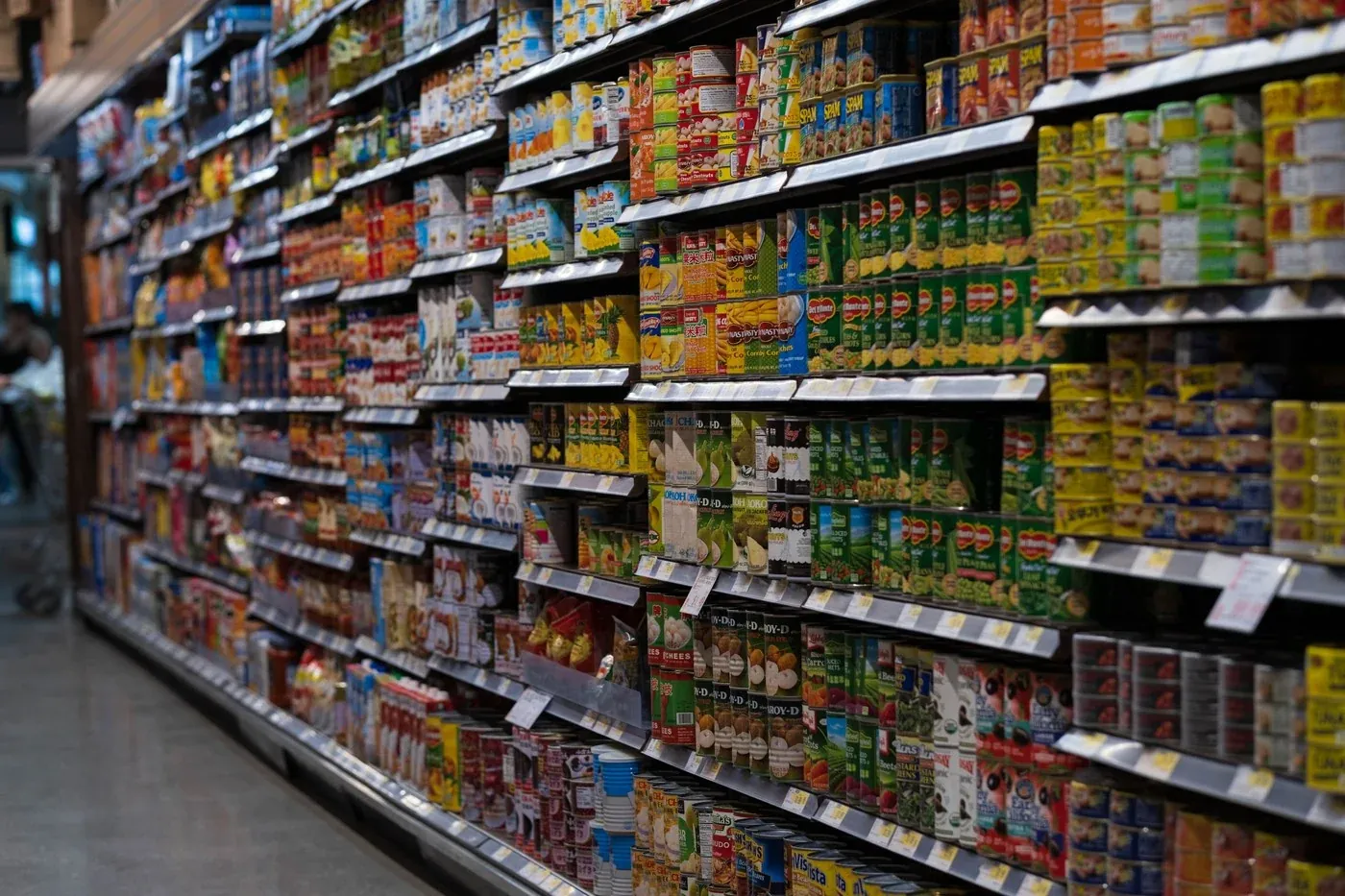 Dry grocery aisle with full canned goods shelving under supermarket track lighting, showing the vertical shelf face that has to hold 300 to 500 lux