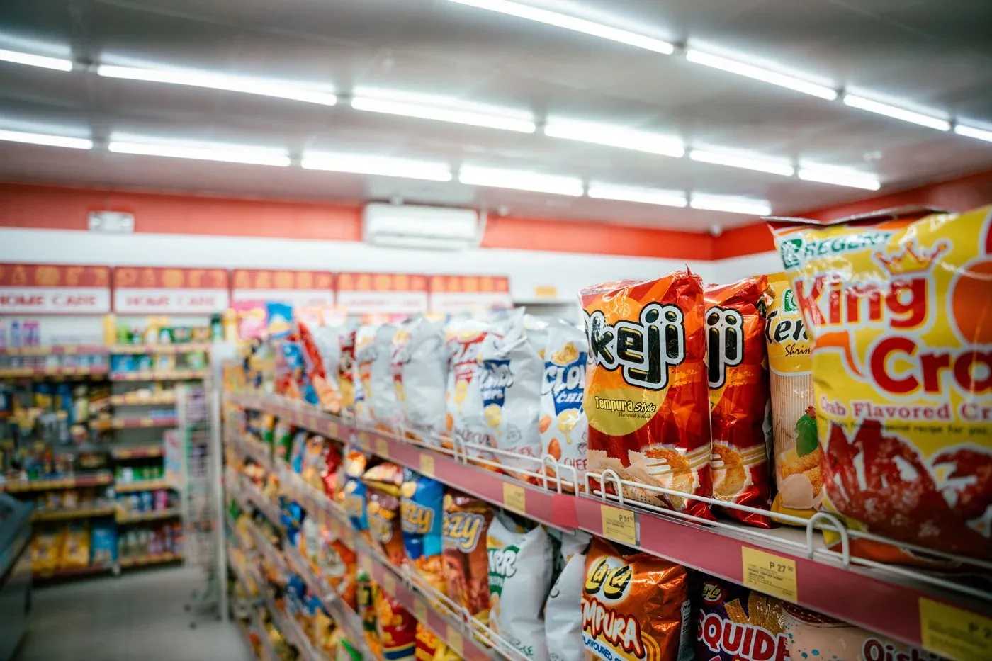 Supermarket snack aisle with crisps and packet goods on both sides under bright track lighting, showing the vertical colour and reds that depend on R9